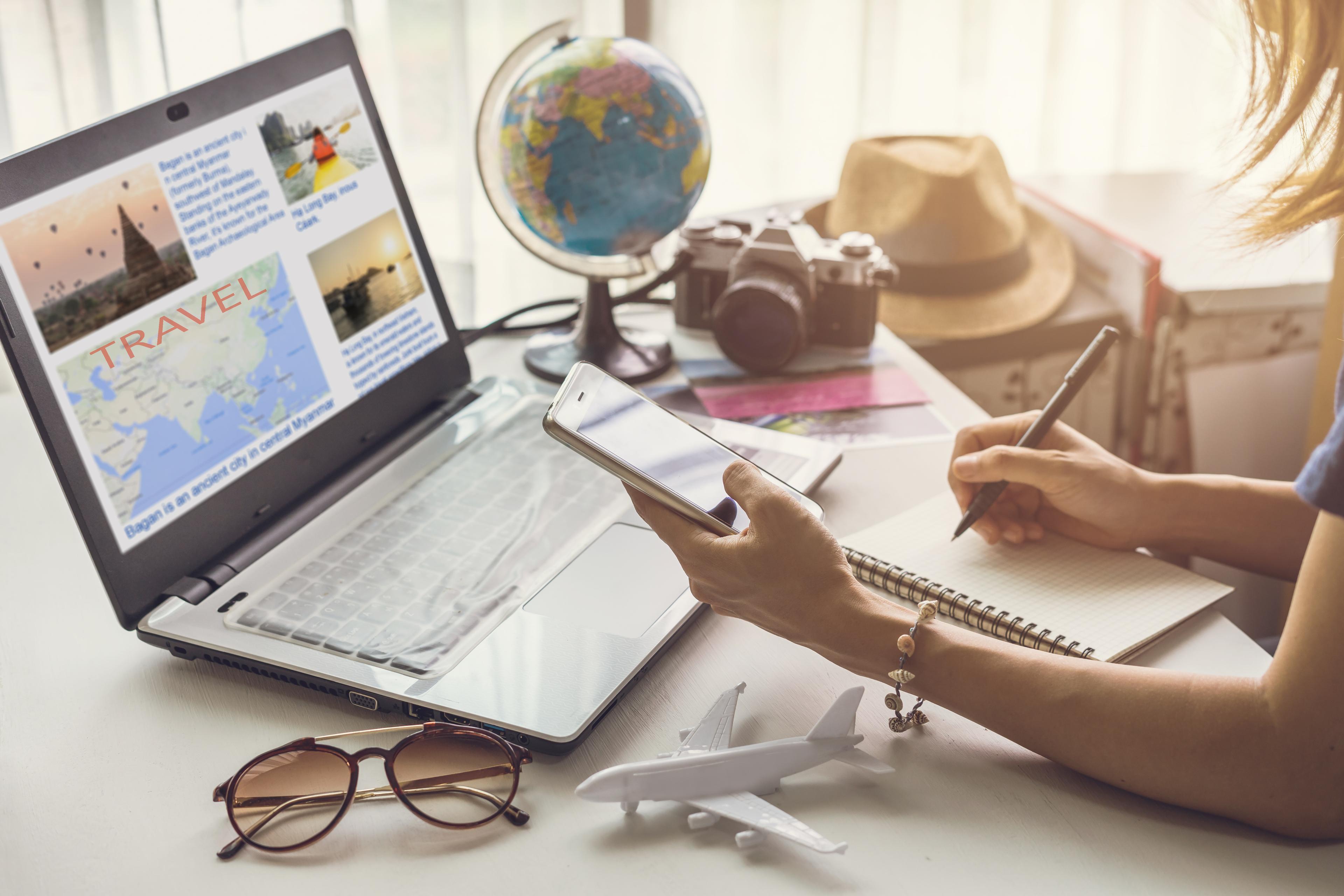 table with laptop saying travel, person holding phone and writing something to notes